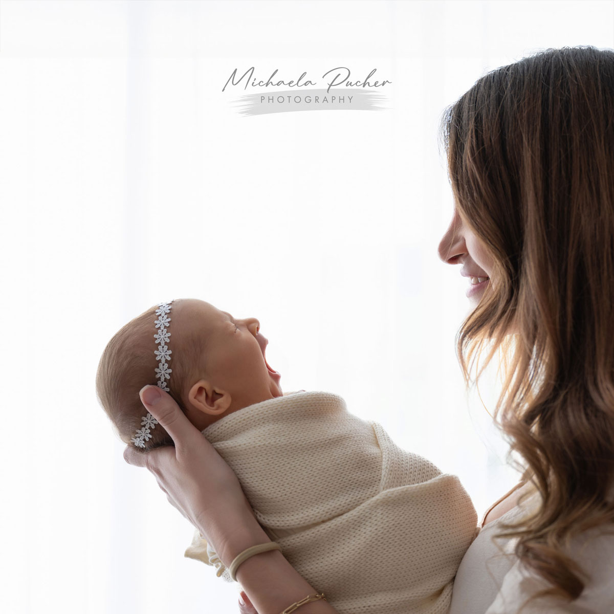 Mama hält ihr eingewickeltes Neugeborenes vor dem hellen Fenster im Arm; das Baby gähnt und die Mutter lächelt es liebevoll an.